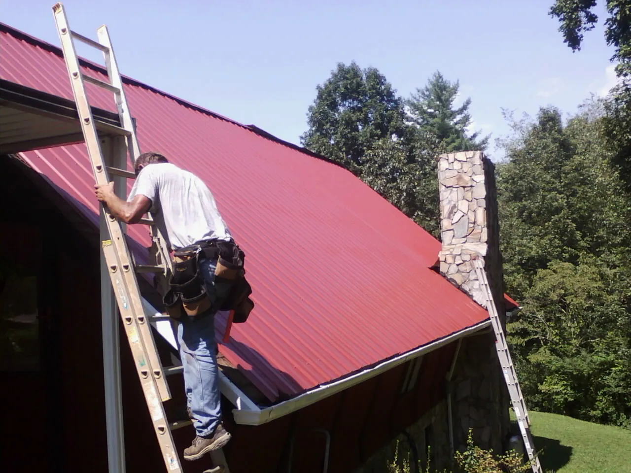 Roofer on a ladder beside a red metal roof and stone chimney — Soffit & Fascia Repair by Roofing Brothers in West Yellowstone